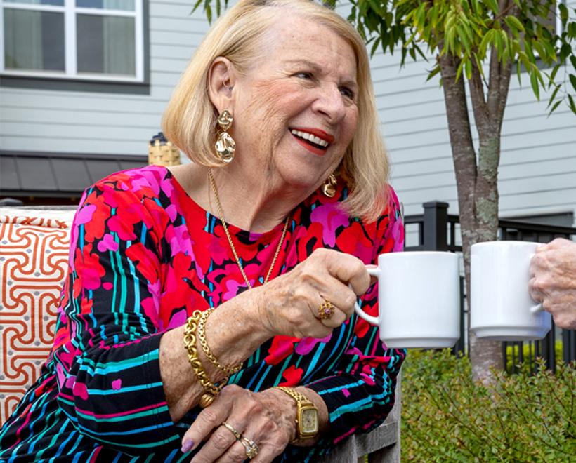 Smiling older woman with blonde hair, wearing a colorful floral top and gold jewelry, holds a white mug while sitting outside, appearing to toast with another person.