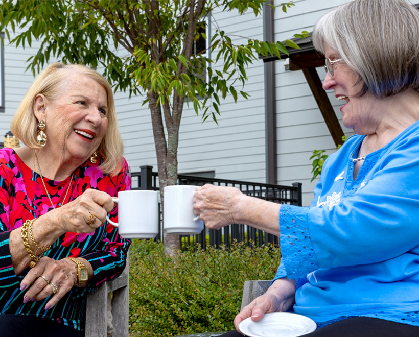 Two older women sit outside on a bench, smiling and clinking white mugs together, enjoying a cheerful conversation on a sunny day with greenery and a building in the background.
