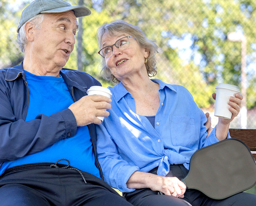 Two older adults sit on a bench outdoors, smiling and talking while holding coffee cups. One holds a pickleball paddle on their lap. Trees and a fence are visible in the background.