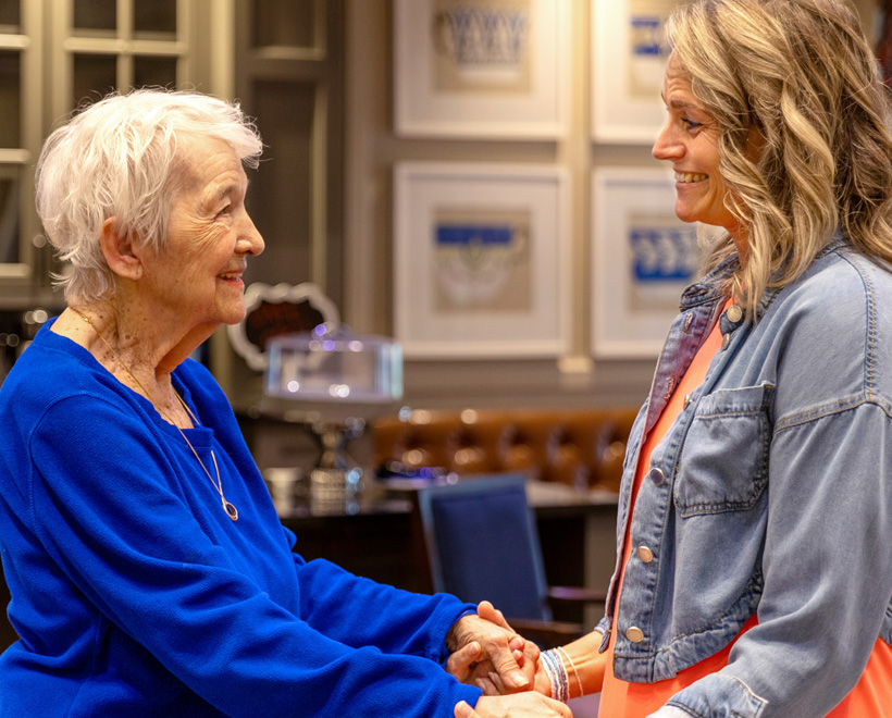 An older woman and a younger woman smile warmly at each other while holding hands indoors. The room has framed artwork on the wall and a cake stand in the background.