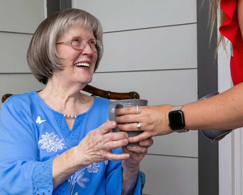 Smiling elderly woman in a blue blouse receives a cup from a person wearing a red shirt and a smartwatch. They are sitting outdoors on a porch.