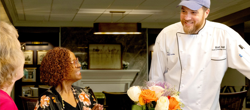 A chef in a white coat and blue cap smiles and talks with two women seated at a table with a bouquet of flowers in a warmly lit restaurant.