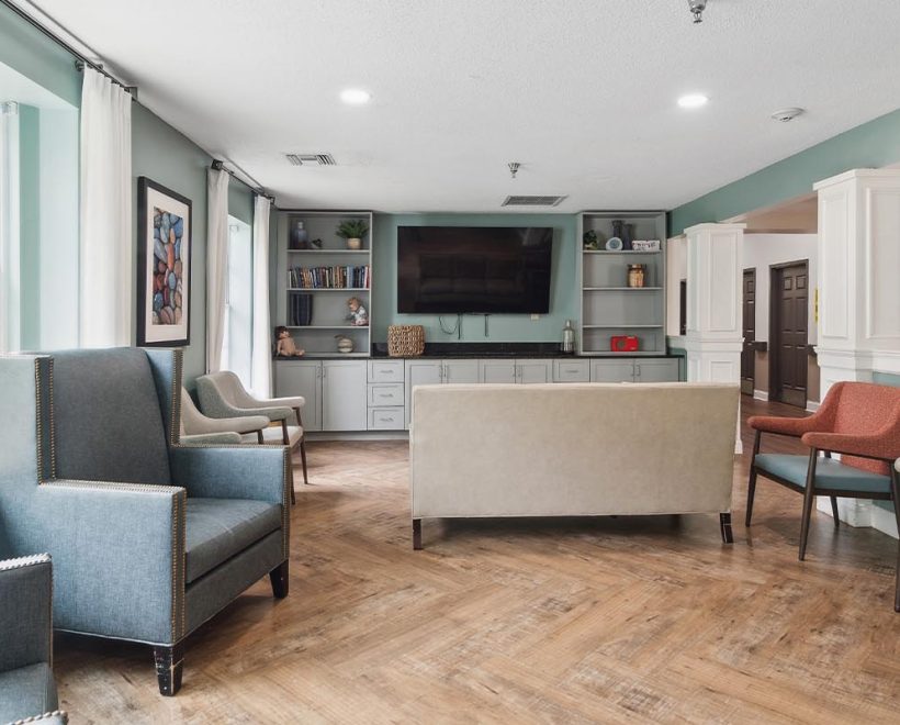 A bright, modern lounge with armchairs, a sofa, wall-mounted TV, bookshelves, and large windows letting in natural light. The room has wood flooring and pastel-colored walls.