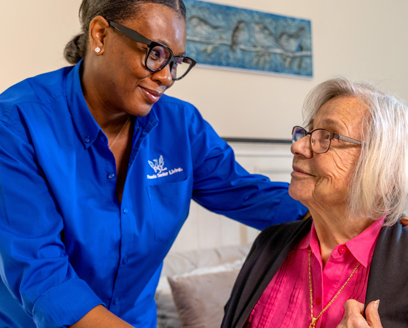 A caregiver in a blue shirt smiles and gently supports an elderly woman with gray hair and glasses, who is seated and smiling back. They appear to share a warm, caring moment in a home setting.