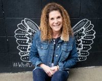 Teri Marinko wearing a denim jacket and jeans, sits on the ground smiling, with black and white illustrated wings on the wall behind them, creating the illusion of having wings.