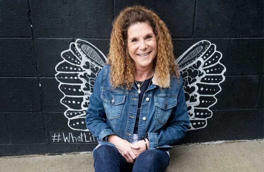 Teri-Marinko - Phoenix Senior Living Teri Marinko wearing a denim jacket and jeans, sits on the ground smiling, with black and white illustrated wings on the wall behind them, creating the illusion of having wings.