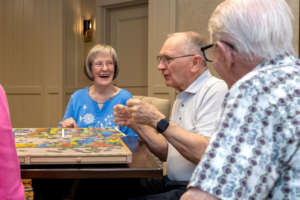 Three older adults sit around a table, working on a colorful jigsaw puzzle. One woman smiles while two men talk. Indoor setting with soft lighting.