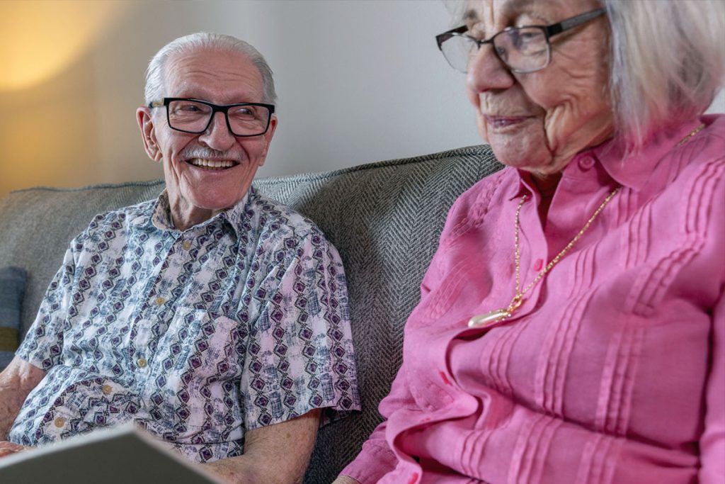 An elderly man and woman sit on a couch together. The man wears glasses and a patterned shirt, smiling at the woman, while she wears glasses and a pink shirt, looking down at something in her hands.