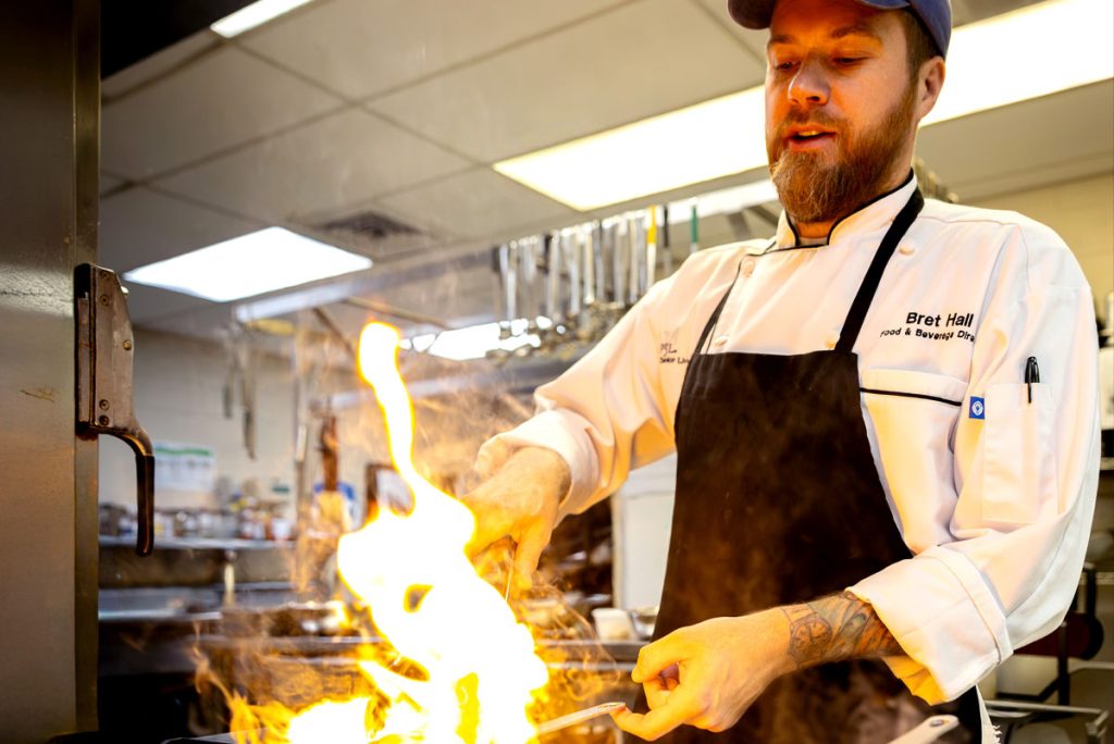 A chef in a white coat and black apron cooks at a stovetop with high flames rising from a pan in a commercial kitchen.