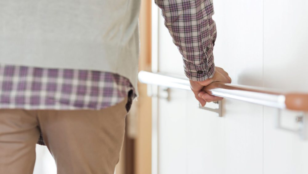 A person wearing a plaid shirt and beige pants holds onto a handrail while walking down a hallway.