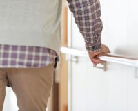 A person wearing a plaid shirt and beige pants holds onto a handrail while walking down a hallway.
