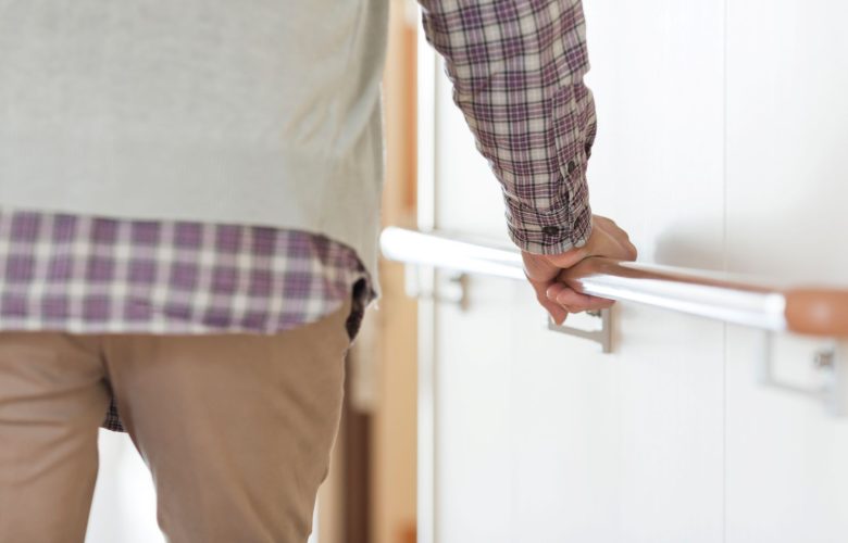 A person wearing a plaid shirt and beige pants holds onto a handrail while walking down a hallway.