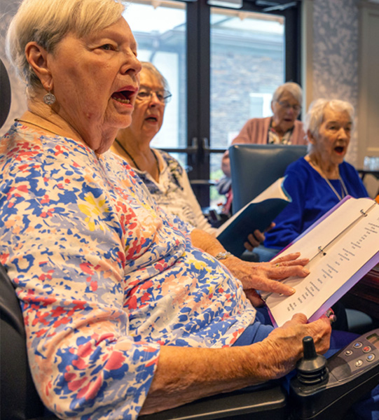 Four elderly women sit together indoors, singing from songbooks. The woman in front wears a floral shirt and holds a binder with lyrics. They appear engaged in a group activity, possibly a choir or singing session.