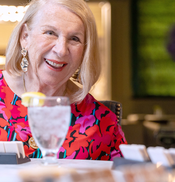 Smiling older woman with blonde hair sits at a table, wearing a colorful red and pink floral top. A glass of water with a lemon slice and several small dishes are in front of her. The background is softly blurred.