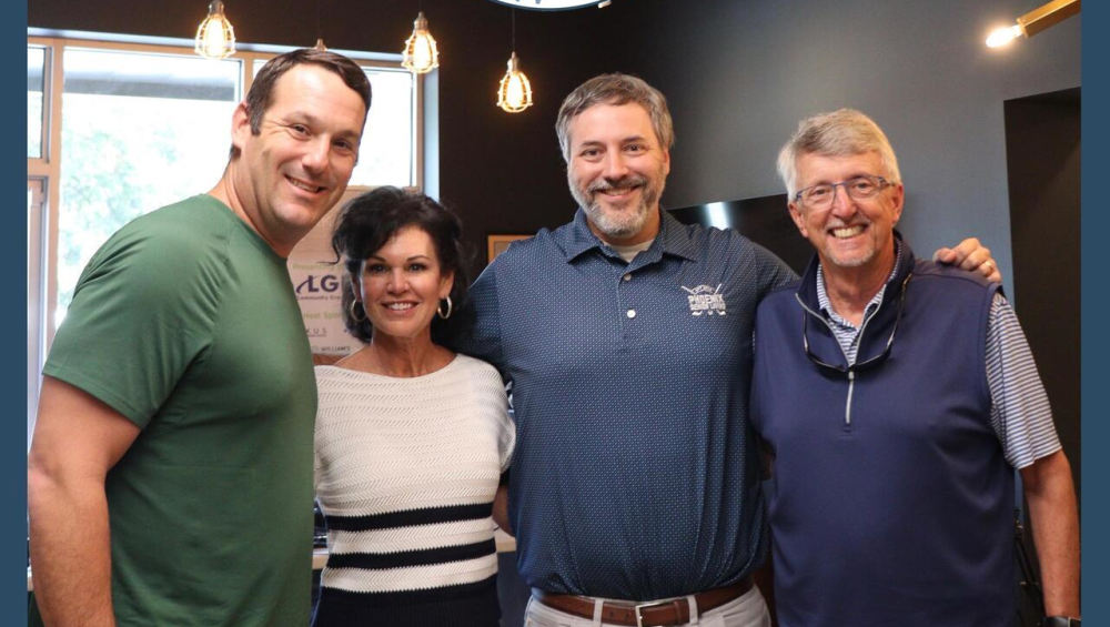 Roswell Brew Talks Photo - Phoenix Senior Living Four people stand smiling with arms around each other beneath a Roswell Brew Talks RBT Podcast sign in a modern, warmly lit room with exposed ceiling beams and hanging lights.