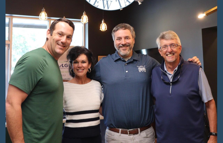 Roswell Brew Talks Photo - Phoenix Senior Living Four people stand smiling with arms around each other beneath a Roswell Brew Talks RBT Podcast sign in a modern, warmly lit room with exposed ceiling beams and hanging lights.