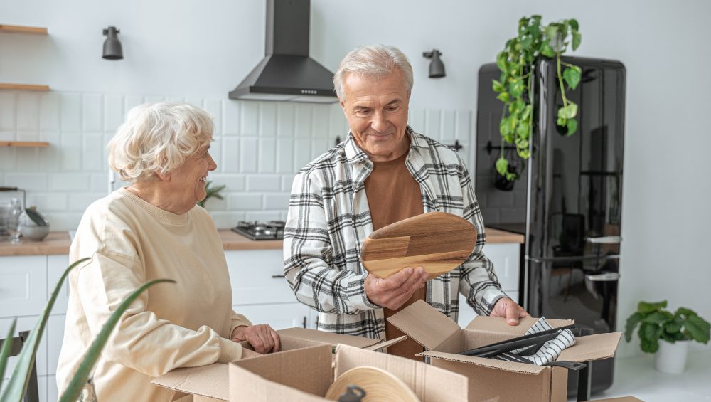 Mature couple standing at the kitchen with moving boxes at the new house - Phoenix Senior Living An older couple unpacks kitchen items from cardboard boxes in their kitchen.