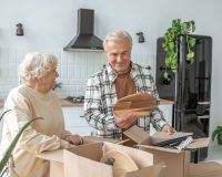 An older couple unpacks kitchen items from cardboard boxes in their kitchen.