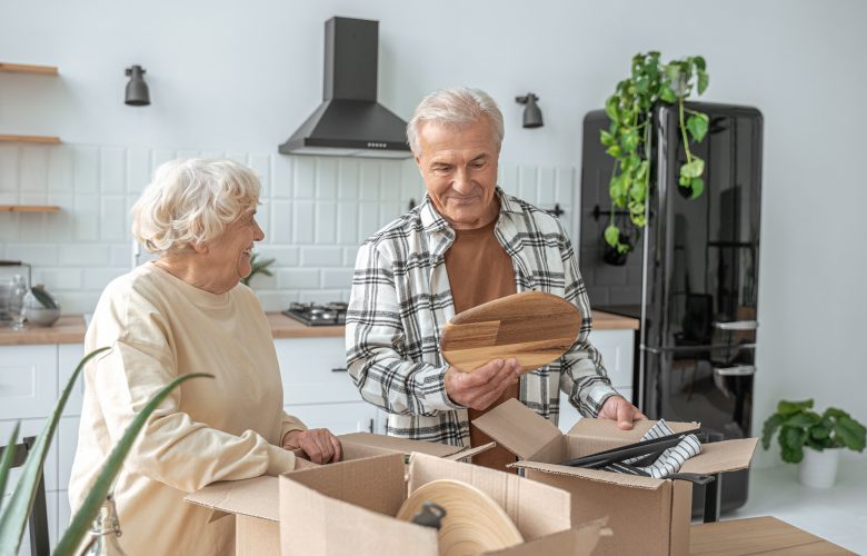 Mature couple standing at the kitchen with moving boxes at the new house - Phoenix Senior Living An older couple unpacks kitchen items from cardboard boxes in their kitchen.