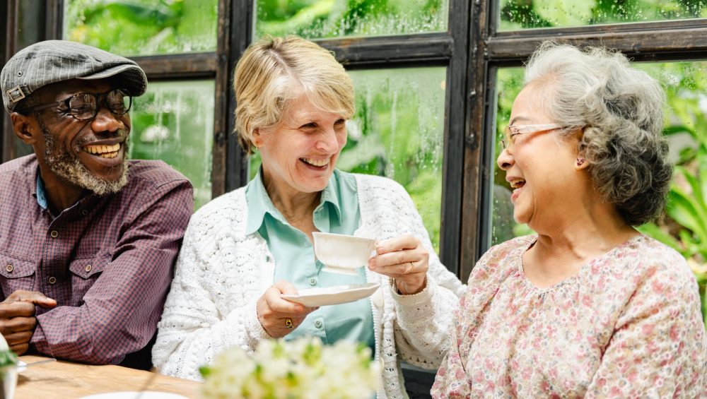PSL three senior friends enjoying tea and laughing together_1226624472 - Phoenix Senior Living Three older adults laughing while sitting at a table with tea in a greenhouse.