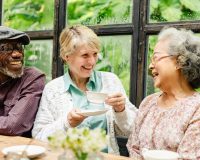 Three older adults laughing while sitting at a table with tea in a greenhouse.