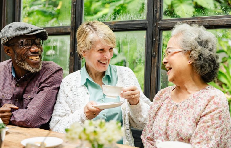 Three older adults laughing while sitting at a table with tea in a greenhouse.
