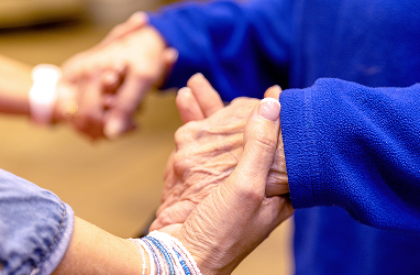 Close-up of two people holding hands, one with older-looking hands wearing a blue sleeve, and the other with younger hands, both clasped in a supportive gesture.