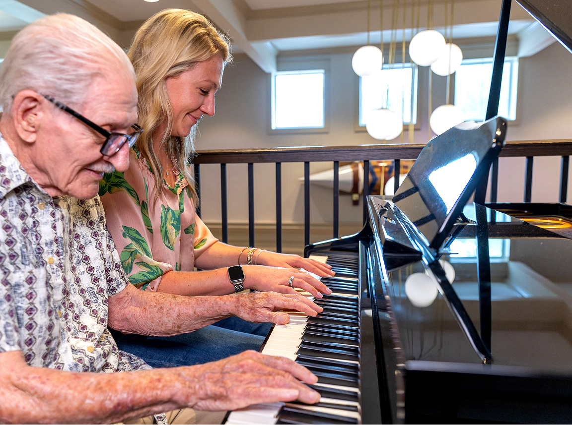 An elderly man and a woman sit together playing a grand piano.