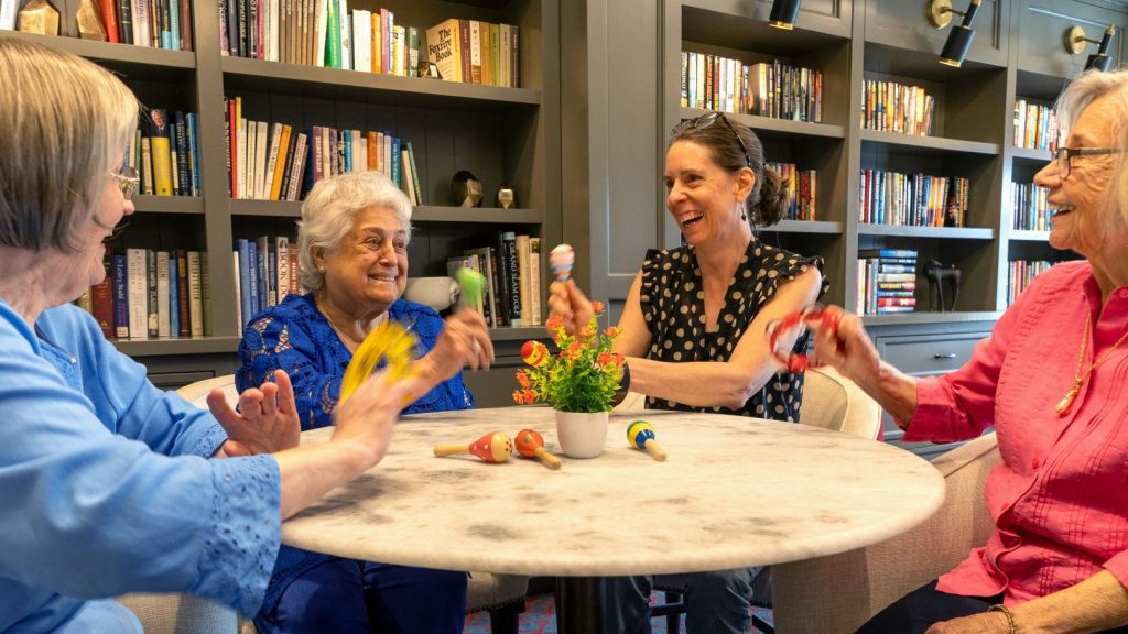 Four women sitting around a round table in a library, smiling and playing with small colorful maracas.